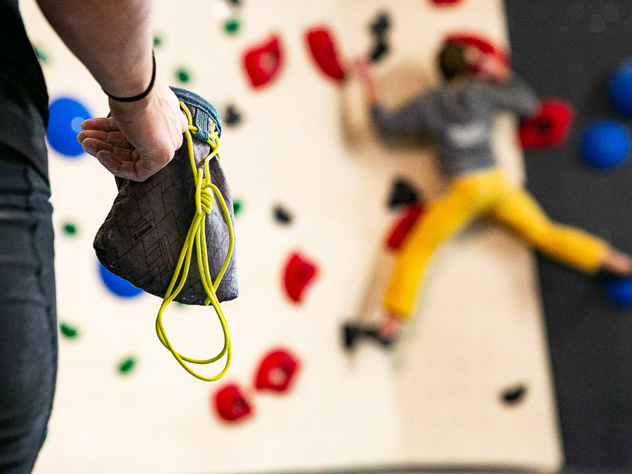 Man sieht eine Hand, die in den Chalk-Beutel greift, um die Hände vor dem Klettern mit Kletter-Chalk einzureiben. Im Hinterhrund sieht man einen Mann, wie er an der Boulderwand ein Tour klettert.