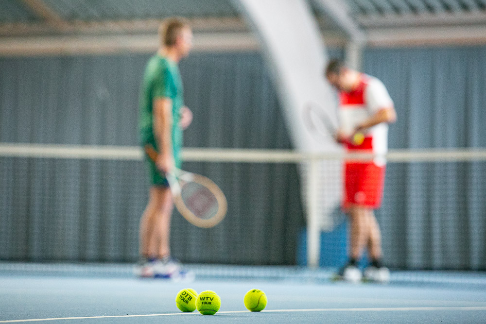 Drei Tennisbälle liegen auf dem Teppich in der Tennishalle. Im Hintergrund sieht man unscharf zwei Personen mit Tennisschlägern am Netz, die sich unterhalten.